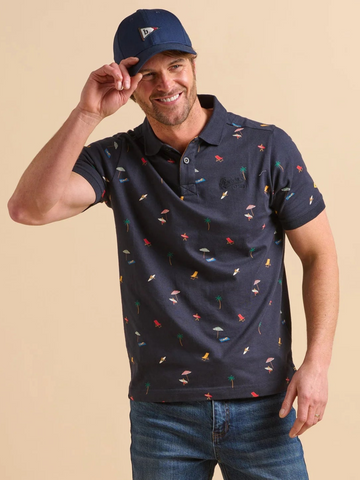 Man wearing a navy patterned polo shirt, blue jeans, and a navy cap on a beige background
